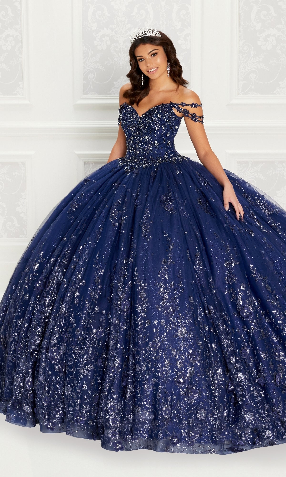 A woman poses gracefully in front of a white ornate wall wearing the Quinceanera Dress PR22145 By Princesa, a sparkling navy blue off-the-shoulder gown with rhinestone embellishments and a corset back.